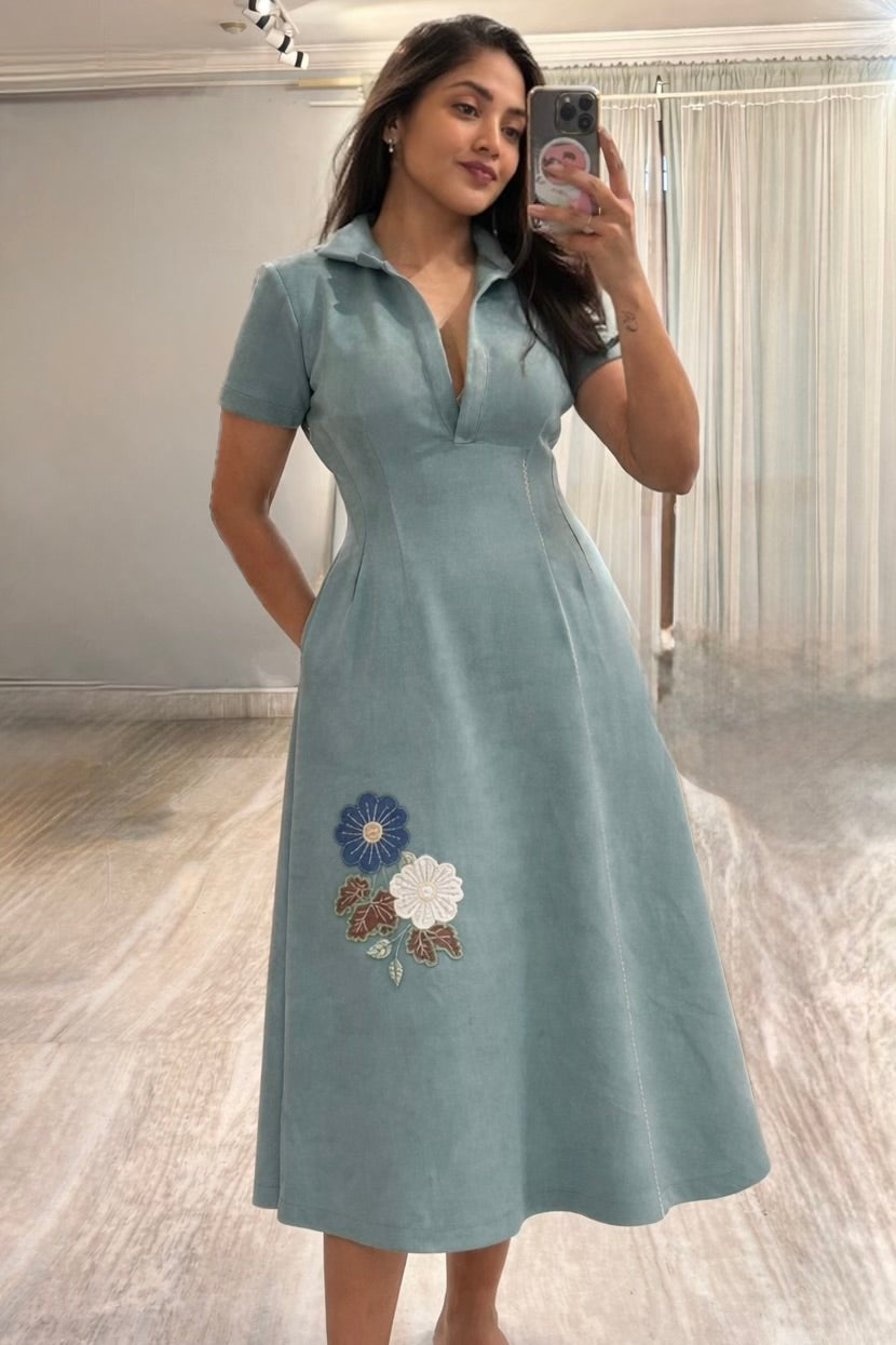 Woman wearing a light blue dress with floral embroidery, taking a mirror selfie.