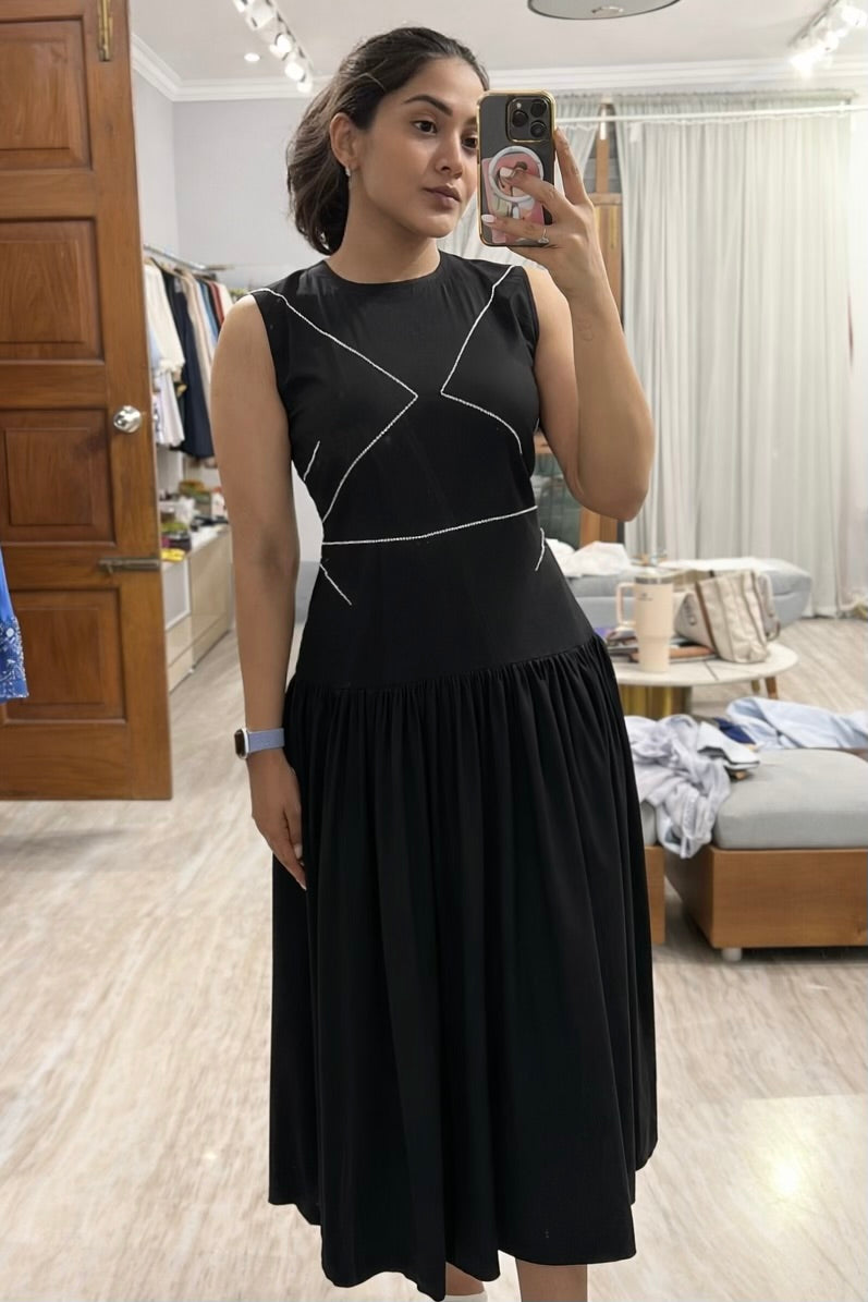 Woman taking a mirror selfie wearing a black dress in a room with wooden flooring and furniture.