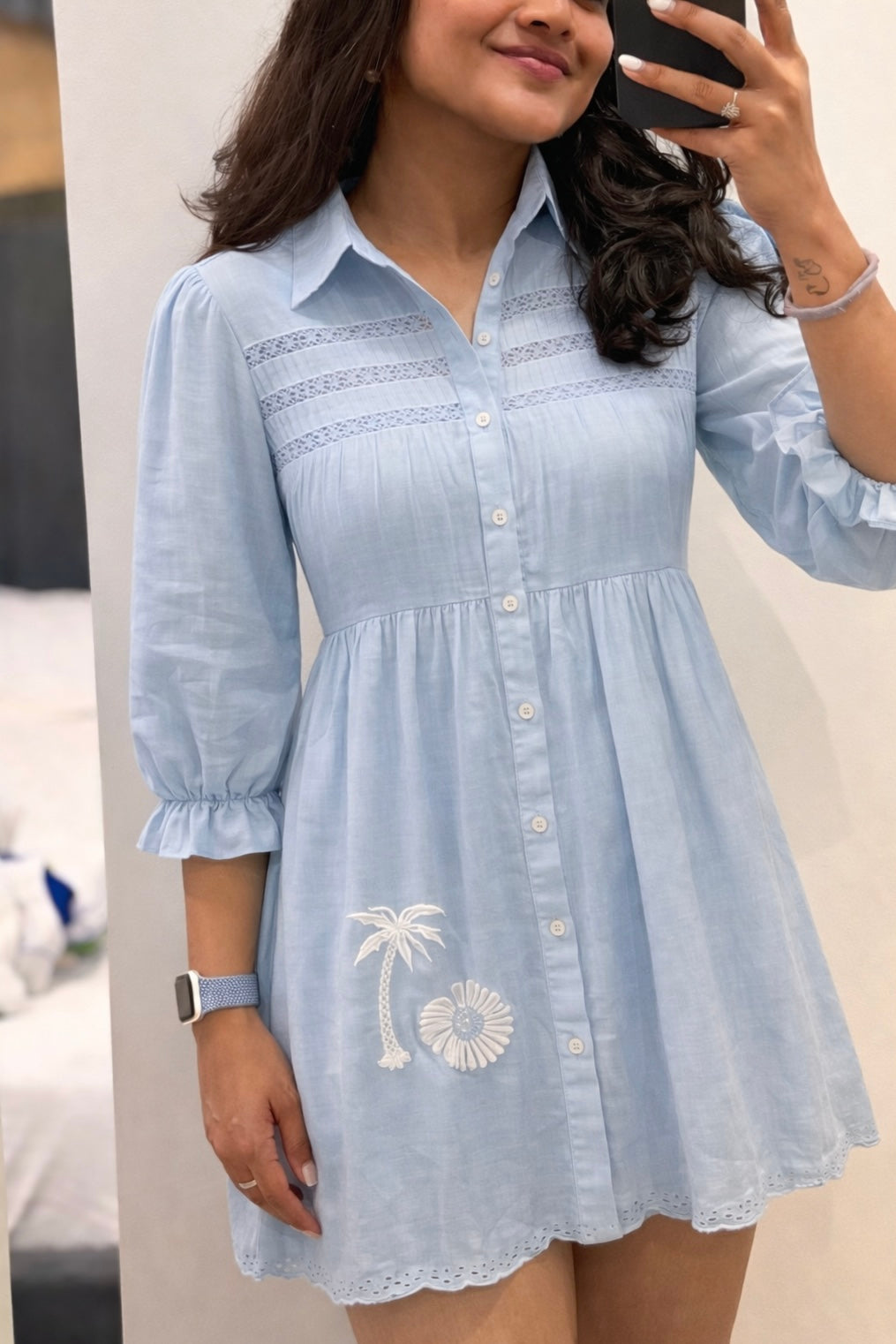 Woman wearing a light blue dress with palm tree design, taking a mirror selfie.