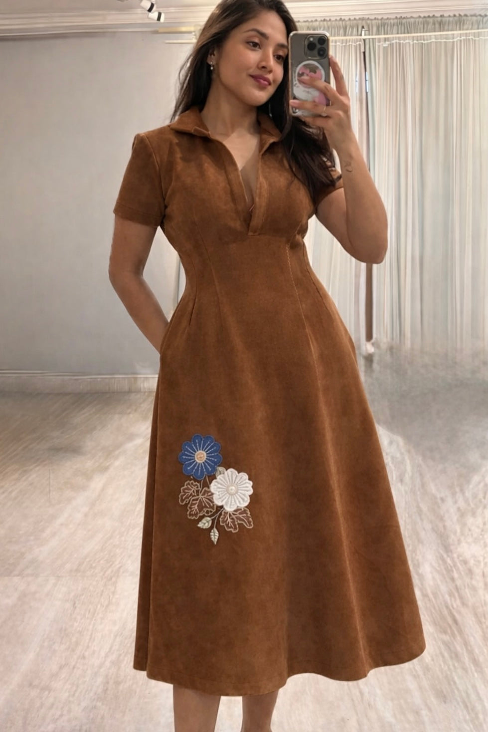 Woman wearing a brown dress with floral embroidery, taking a mirror selfie.