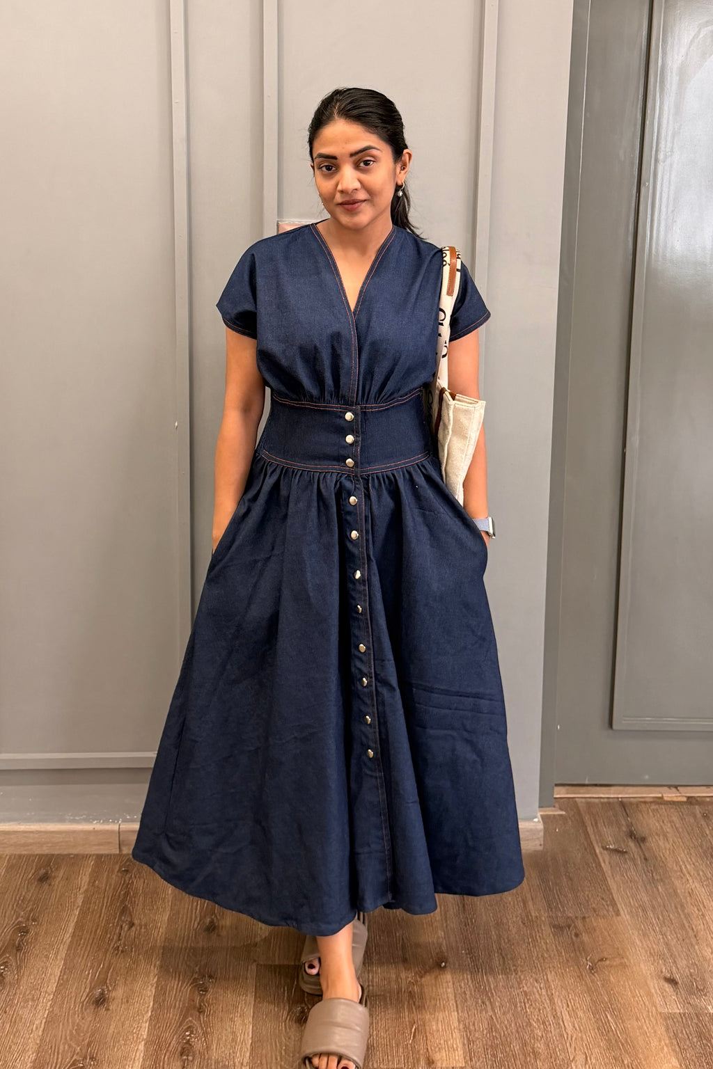 Woman wearing a blue dress standing in a room with gray walls and wooden flooring.