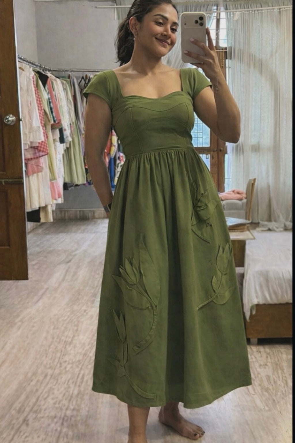 Woman taking a mirror selfie wearing a green dress in a room with clothes and furniture.