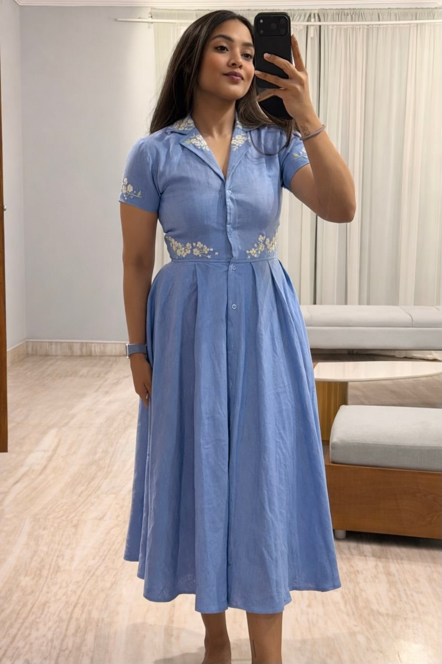 Woman taking a mirror selfie wearing a blue dress with white embroidery in a room.